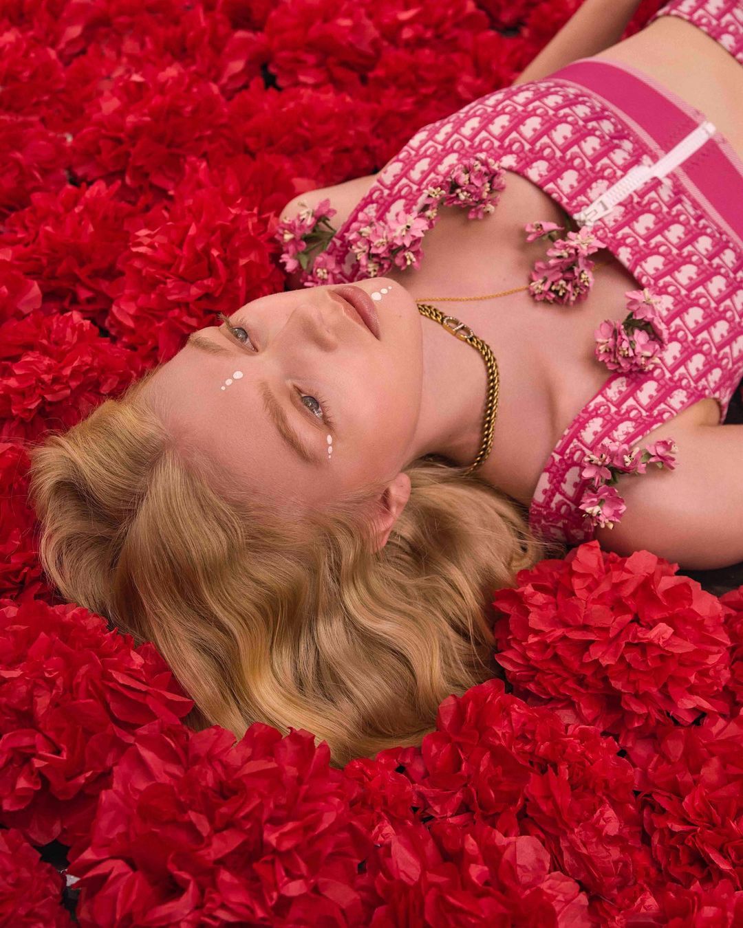 An adult female model lies surrounded by red flowers, wearing a pink patterned top, a gold chain, and white dots under her eyes.