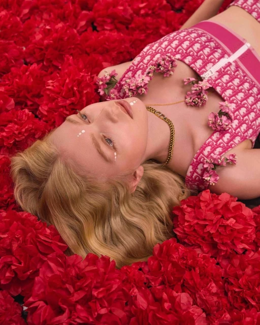 An adult female model lies surrounded by red flowers, wearing a pink patterned top, a gold chain, and white dots under her eyes.