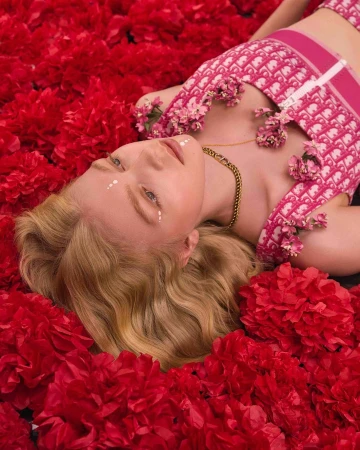 An adult female model lies surrounded by red flowers, wearing a pink patterned top, a gold chain, and white dots under her eyes.