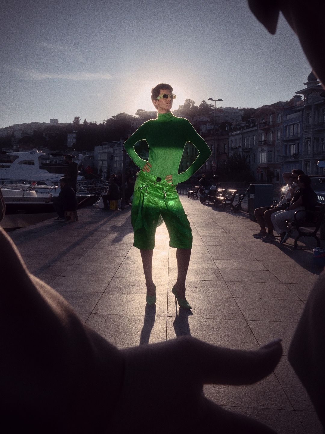 Fashion model poses in green outfit on city street with waterfront background, framed by blurry hands, bathed in sunny, bright light.
