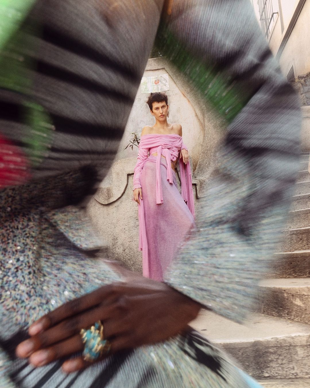 Fashion shot of a woman in a pink outfit leaning against a weathered staircase with an obscured foreground person wearing an ornate ring.
