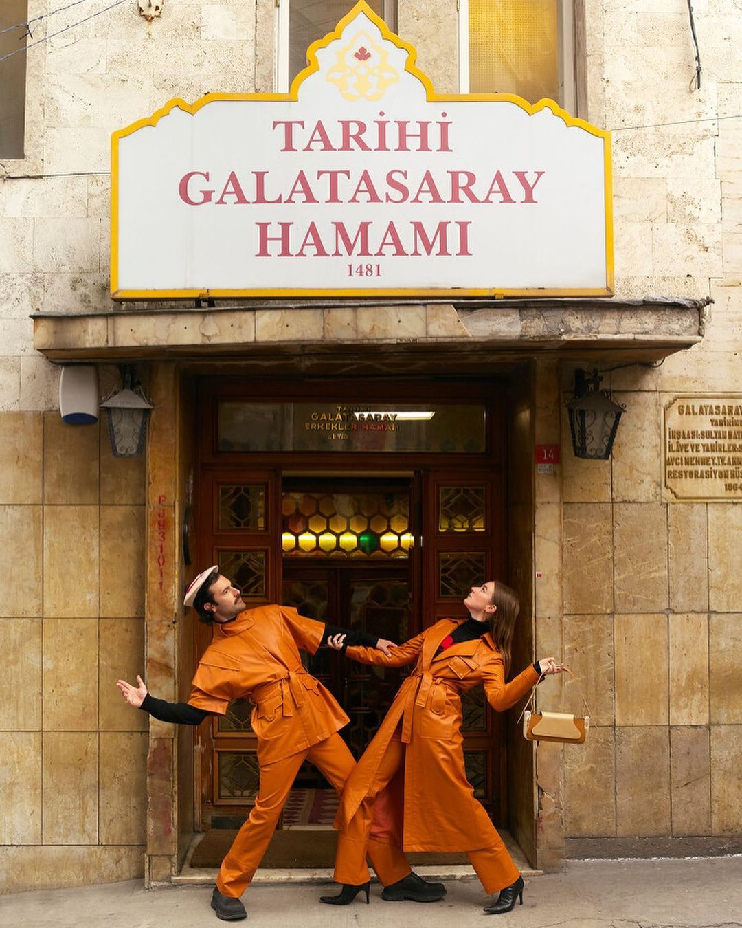 A man and woman in matching orange outfits strike a pose in front of the Tarihi Galatasaray Hamami building in Istanbul, Turkey.