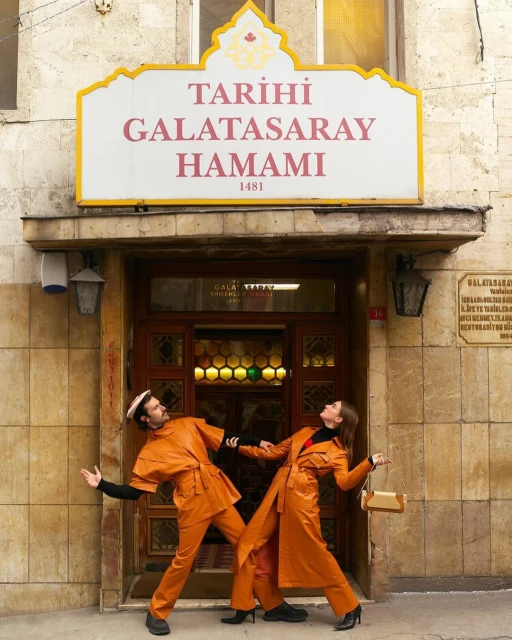 A man and woman in matching orange outfits strike a pose in front of the Tarihi Galatasaray Hamami building in Istanbul, Turkey.