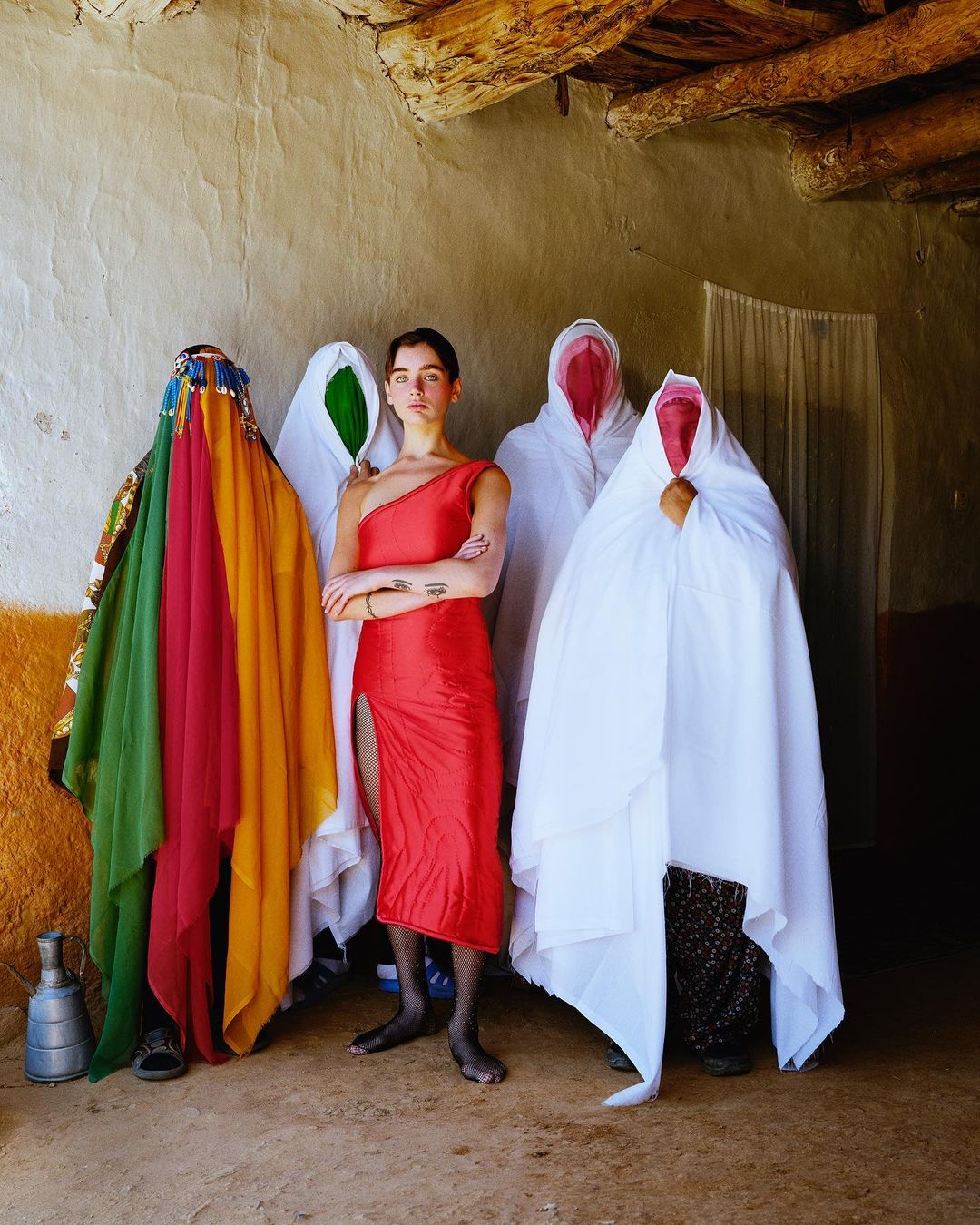A woman in a red dress poses in front of a group of people shrouded in white sheets, one woman stands out in colorful traditional dress.
