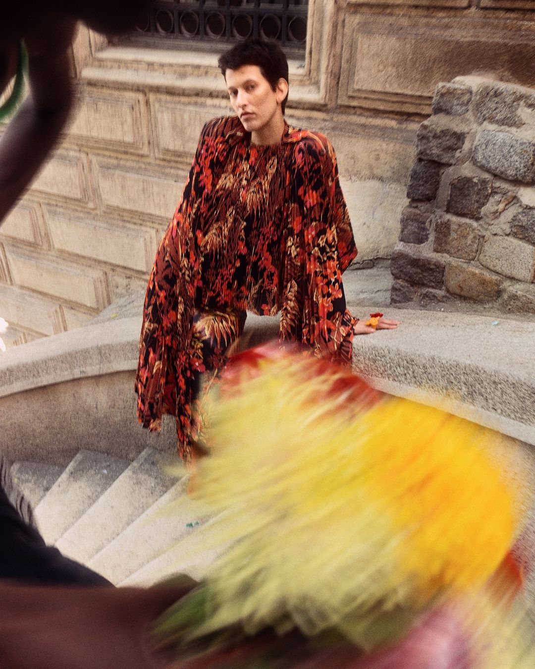 Woman in patterned top posing on stairs outside a building, captured in a medium shot with motion blur in the foreground.