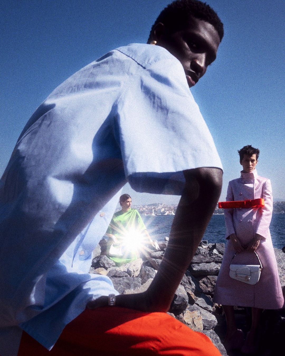 Three fashion models pose on a rocky outcrop, with the sea, city and blue sky in the background on a sunny day in an outdoor setting.