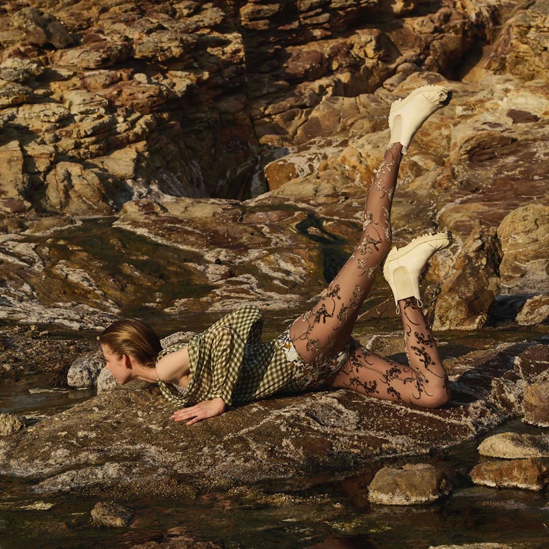 A young female adult in a green plaid top and patterned tights poses on rocks by a body of water in a stylish outdoor fashion shot.