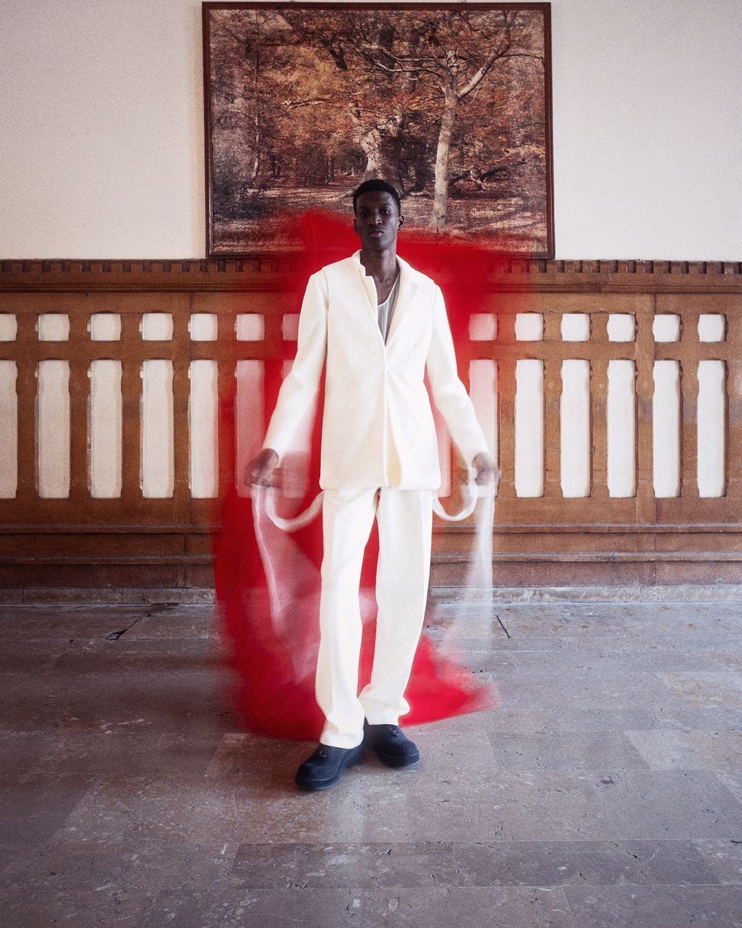 An African male model in a cream suit poses in a bright interior space with a blurred red motion graphic beside him, and a landscape painting.