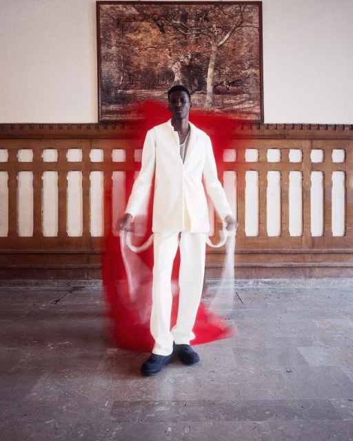 An African male model in a cream suit poses in a bright interior space with a blurred red motion graphic beside him, and a landscape painting.