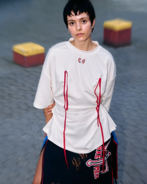 A woman with short black hair poses in a white t-shirt with red details and a patterned skirt in a blurry urban environment.