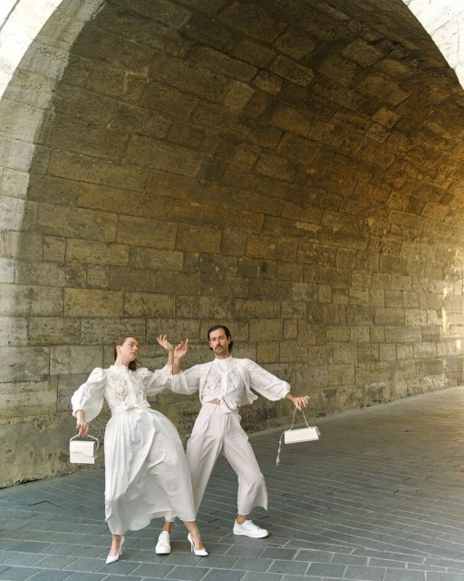 Two fashion models in white outfits and carrying purses pose in a stone archway, creating a symmetrical and stylish composition.