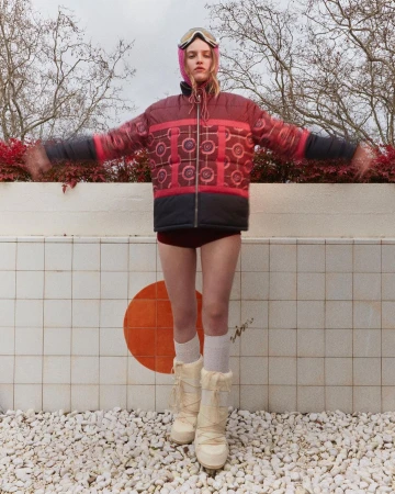Woman in a pink balaclava and puffer jacket poses in front of a tiled wall near white stones and bare trees in an outdoor setting.