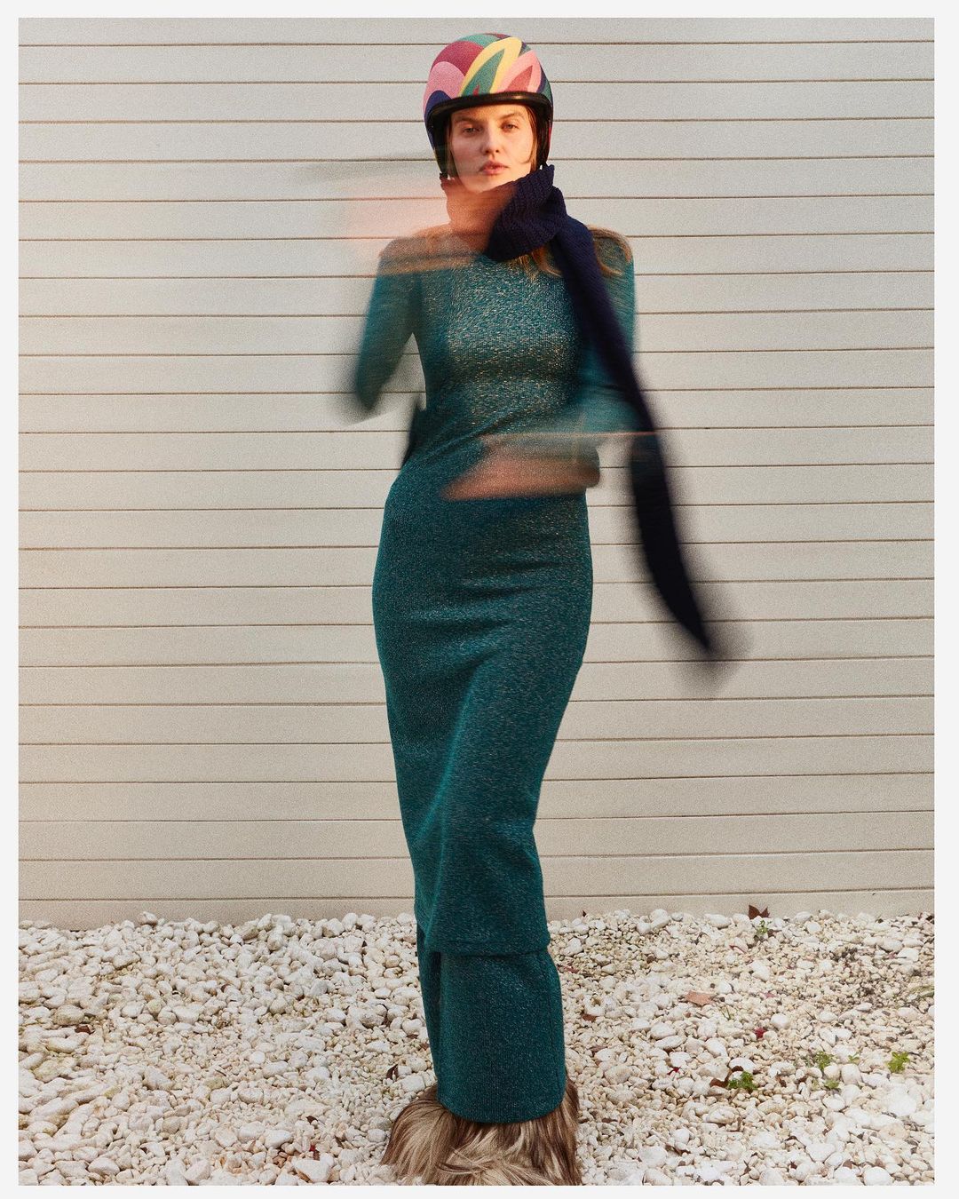 Adult woman in a form-fitting turquoise dress and colorful helmet poses in front of a light-colored wall, creating a fashion image.