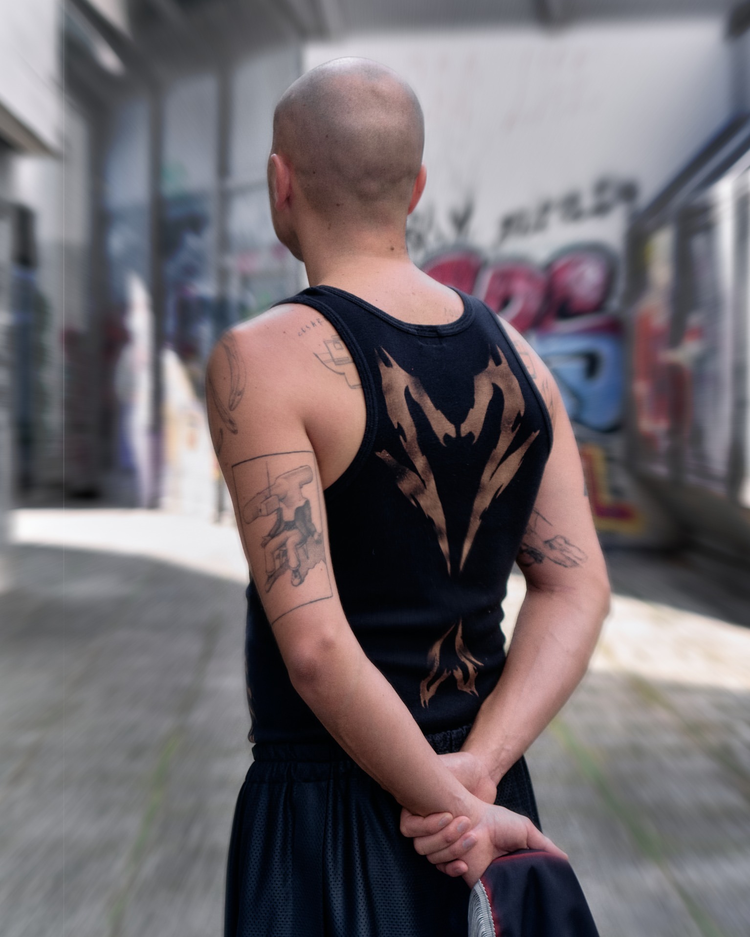 A bald adult male stands in an alleyway, his back to the camera. He has tattoos on his arms and is wearing a tank top.