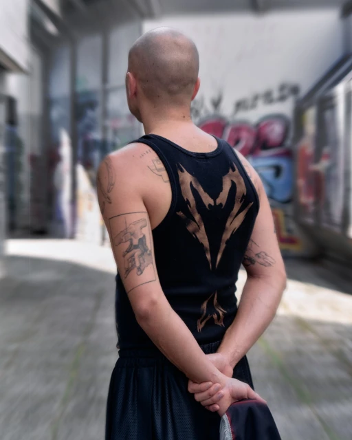 A bald adult male stands in an alleyway, his back to the camera. He has tattoos on his arms and is wearing a tank top.