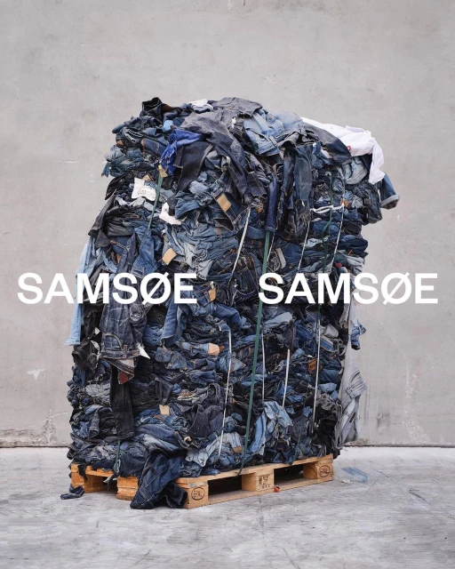 A large bale of recycled denim and clothing sits on a wooden pallet, banded with white tape, in front of a plain concrete wall indoors.