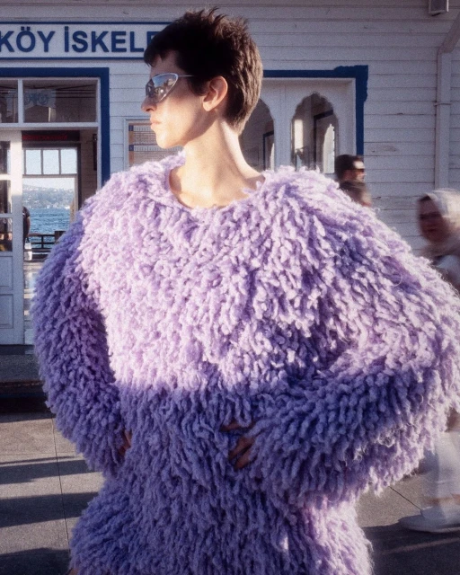 Woman in a fluffy, lavender outfit poses in front of a ferry terminal, wearing sunglasses with blurred people in the background.