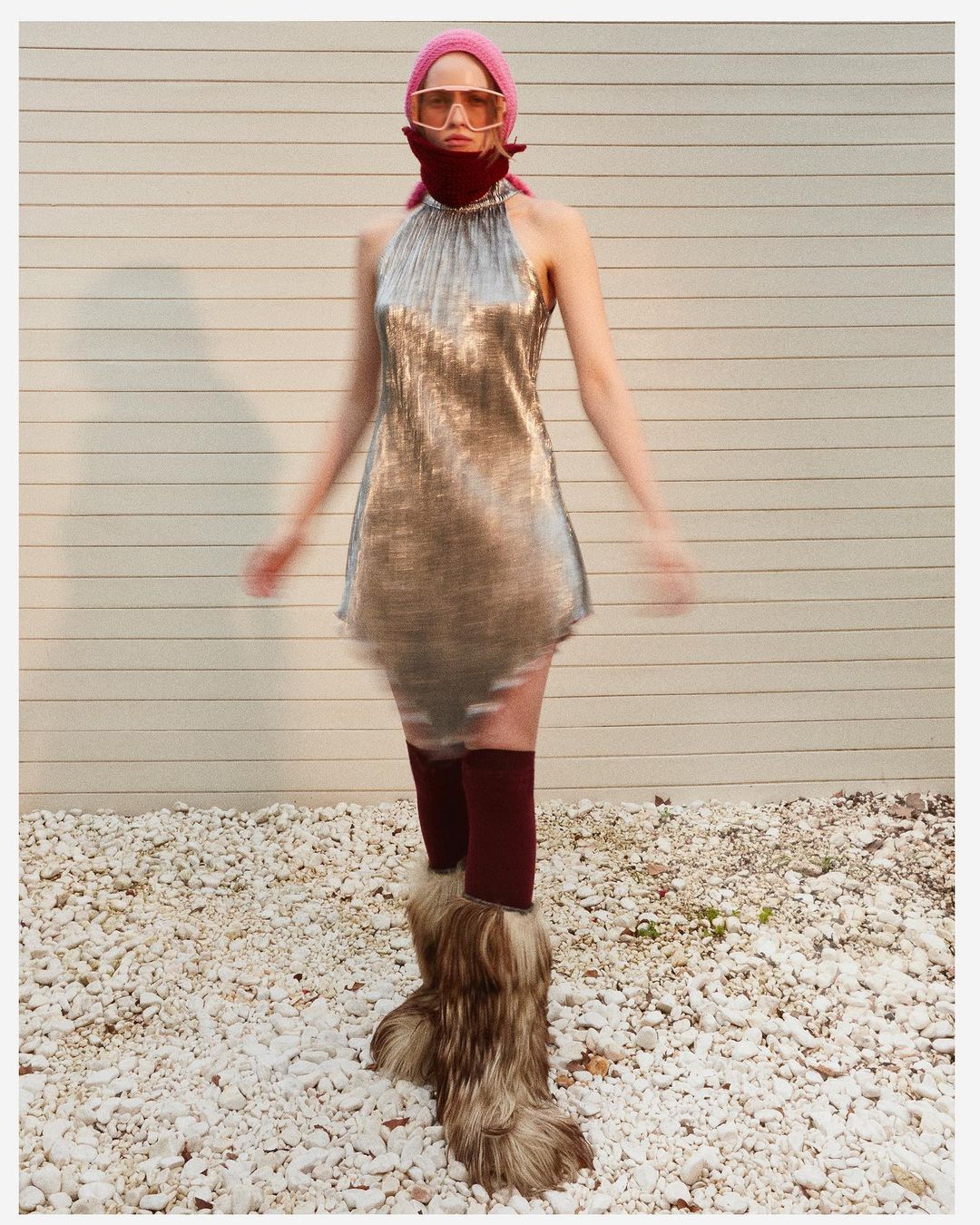 An adult woman wears a shiny silver dress, sunglasses, and furry boots while standing on a gravel surface with a wall in the background.
