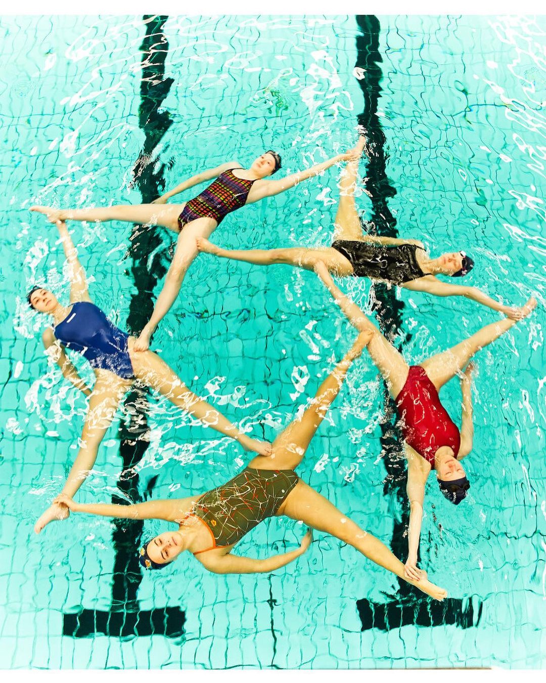 Synchronized swimming team forms star in pool. Six women wear swimsuits and caps, performing in turquoise water with pool tiling.