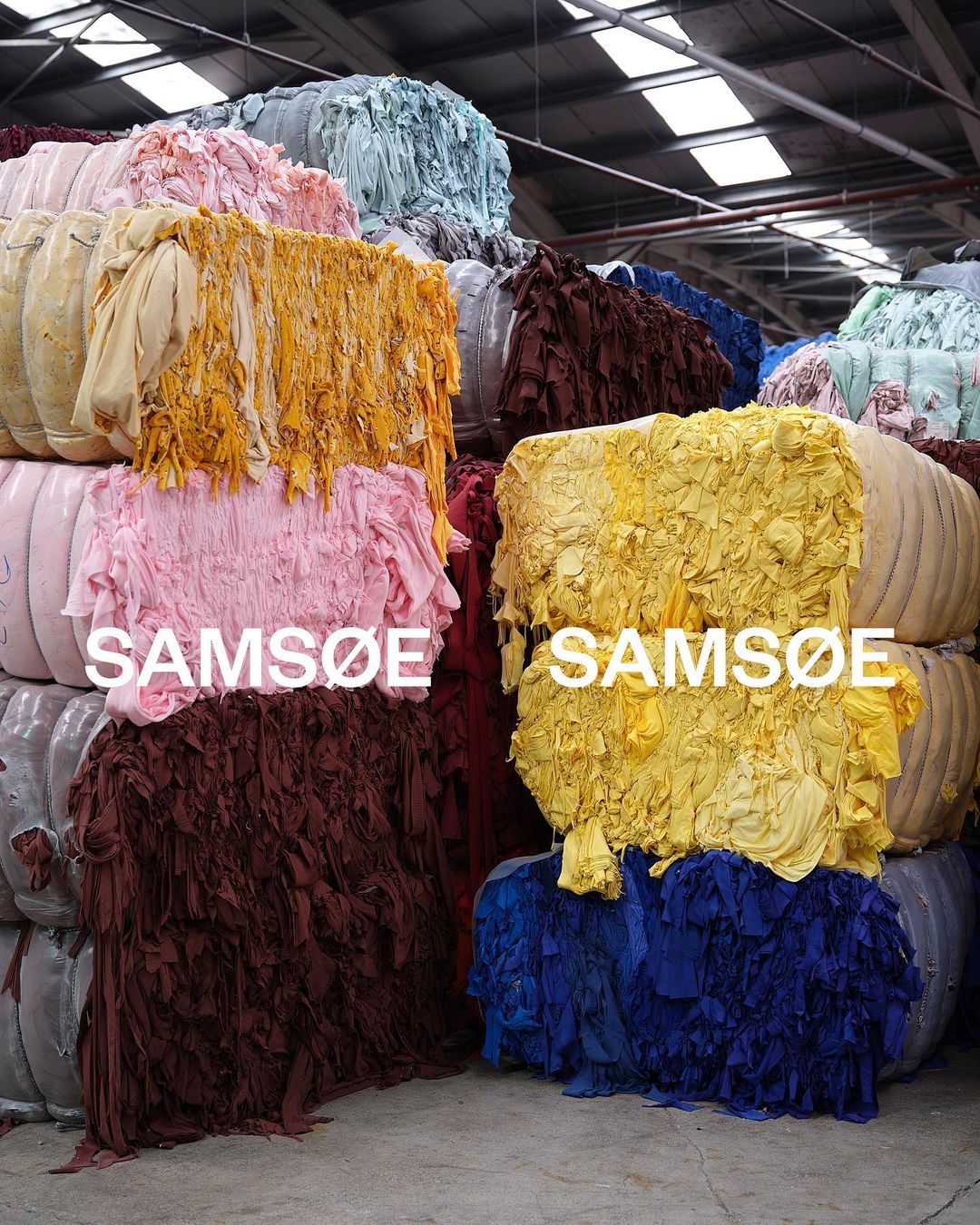 Stacked bales of colorful textile scraps in a warehouse, including pink, yellow, brown, blue, tan, and light blue shades.