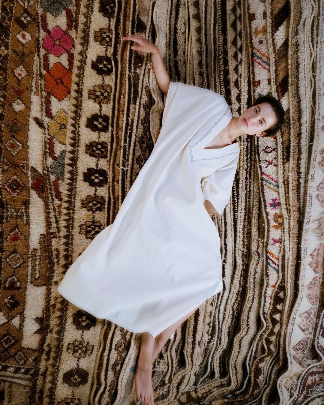 Overhead shot of a woman in a white dress lying on an ornate, patterned rug with outstretched arms. Earth-tone background, fashion editorial.