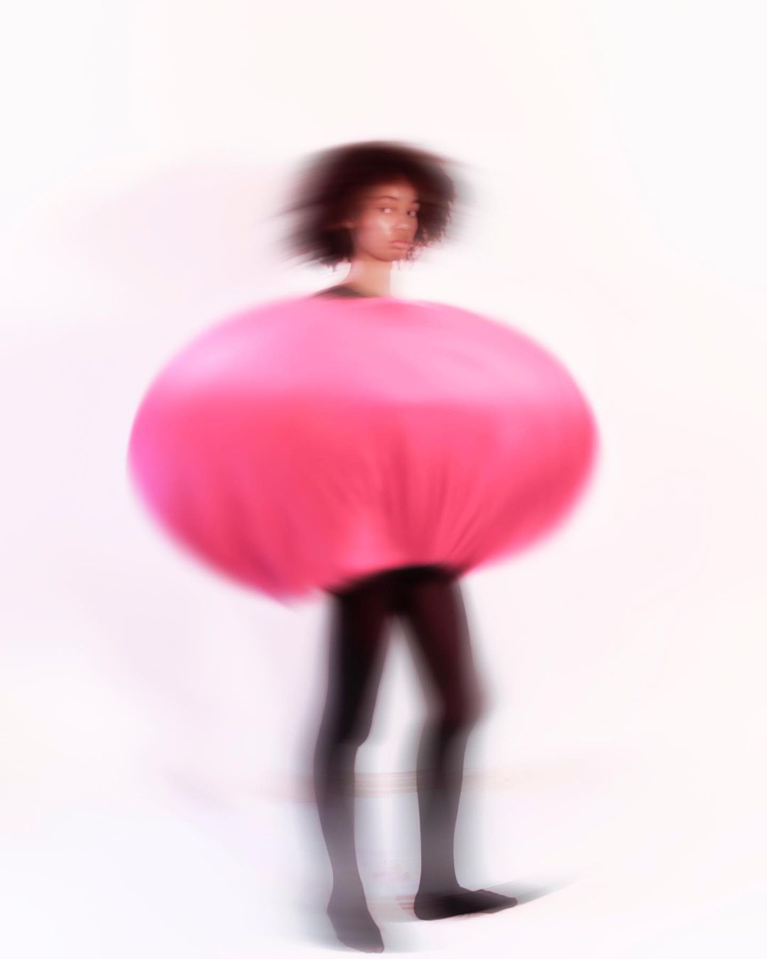 Woman in vibrant pink round avant-garde fashion piece with black leggings against a bright white backdrop in soft focus.