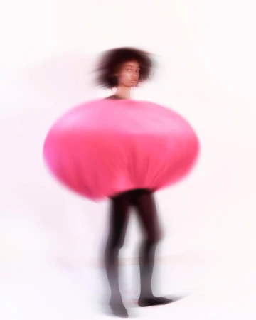 Woman in vibrant pink round avant-garde fashion piece with black leggings against a bright white backdrop in soft focus.