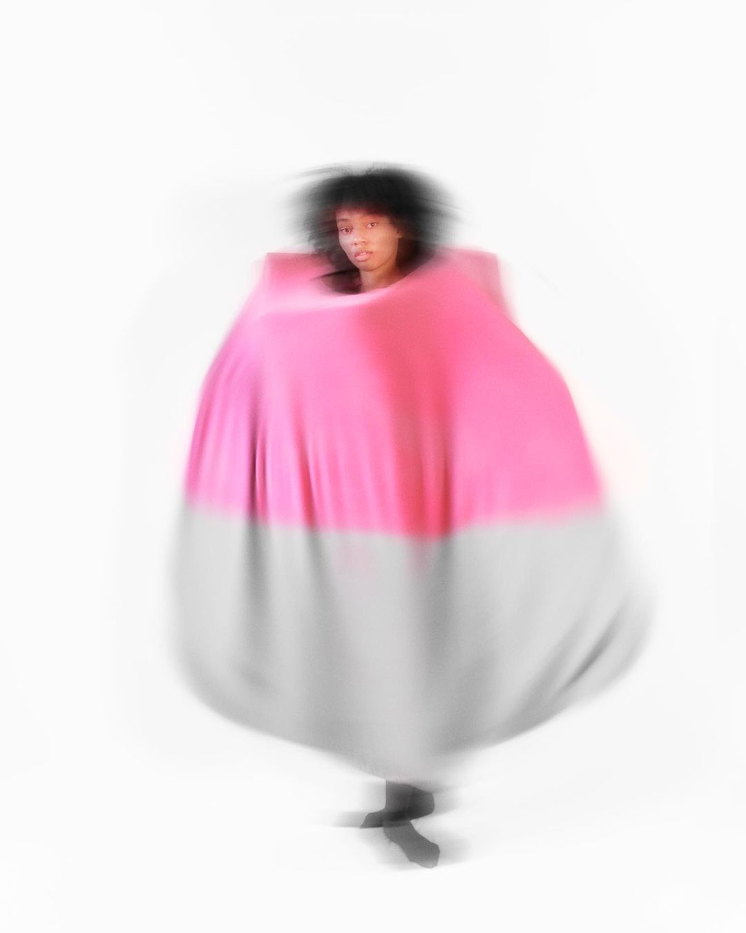 Motion blurred image of a person in a voluminous pink and grey dress against a white backdrop, creating an abstract and dynamic fashion image.