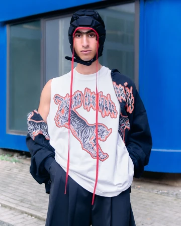 Young man in an oversized tee with a tiger graphic, and a black padded cap poses against a bright blue outdoor wall.
