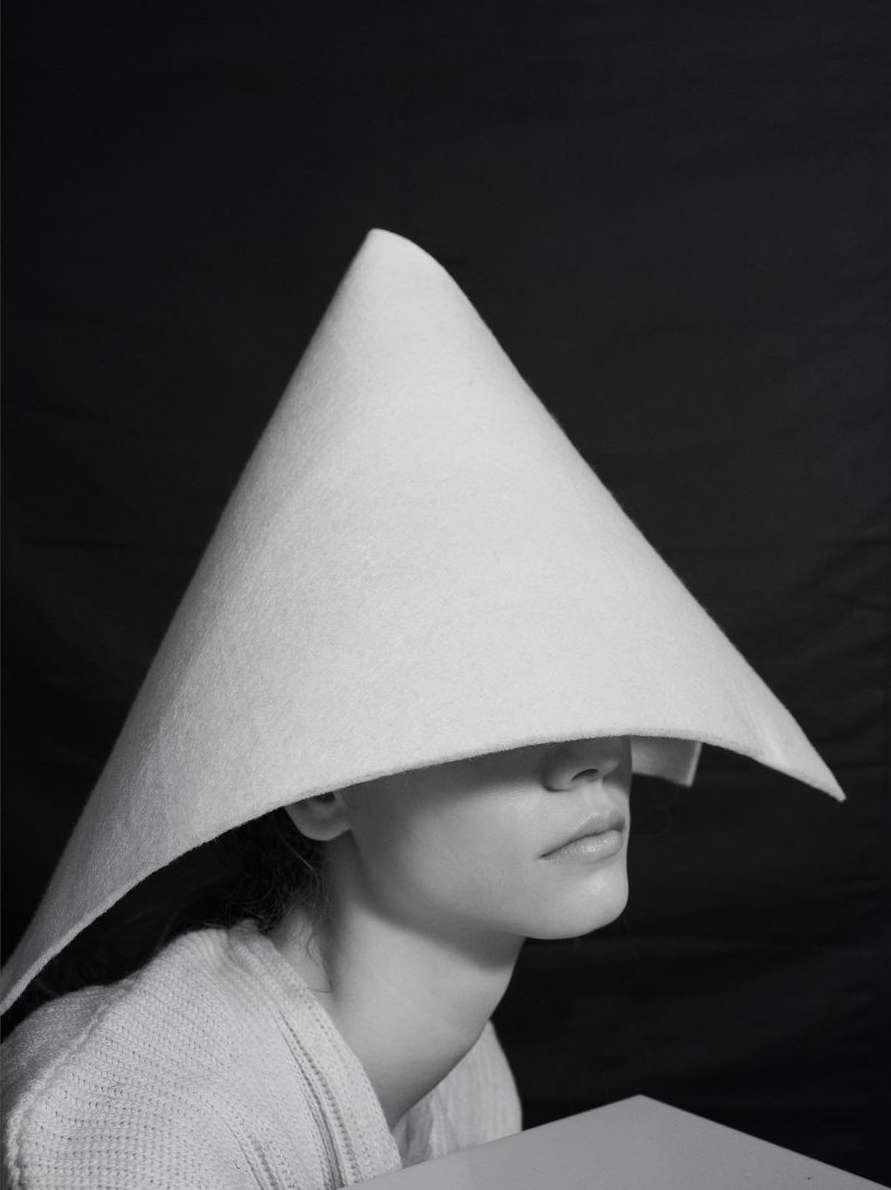 Monochrome portrait of a person wearing a large, cone-shaped white hat covering their eyes, with a black backdrop and minimalist setting.