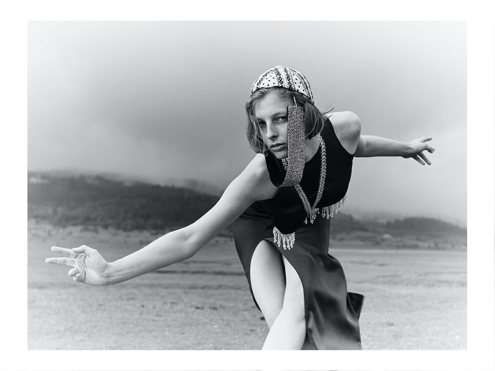 Monochrome fashion portrait of a woman wearing an embellished hat and jewelry, posing outdoors in a dress with a thigh-high slit.