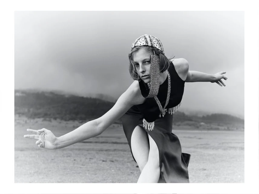 Monochrome fashion portrait of a woman wearing an embellished hat and jewelry, posing outdoors in a dress with a thigh-high slit.