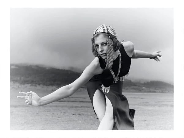 Monochrome fashion portrait of a woman wearing an embellished hat and jewelry, posing outdoors in a dress with a thigh-high slit.