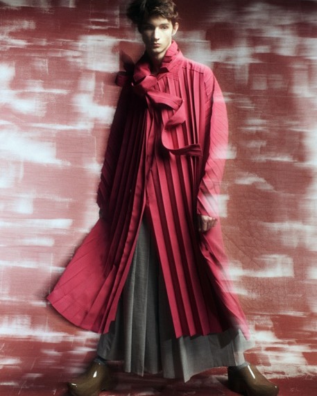 Person posing in a red, pleated coat with unique shoes against a textured background, fashion and style showcased with elegance.