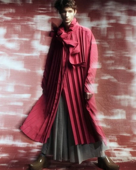 Person posing in a red, pleated coat with unique shoes against a textured background, fashion and style showcased with elegance.