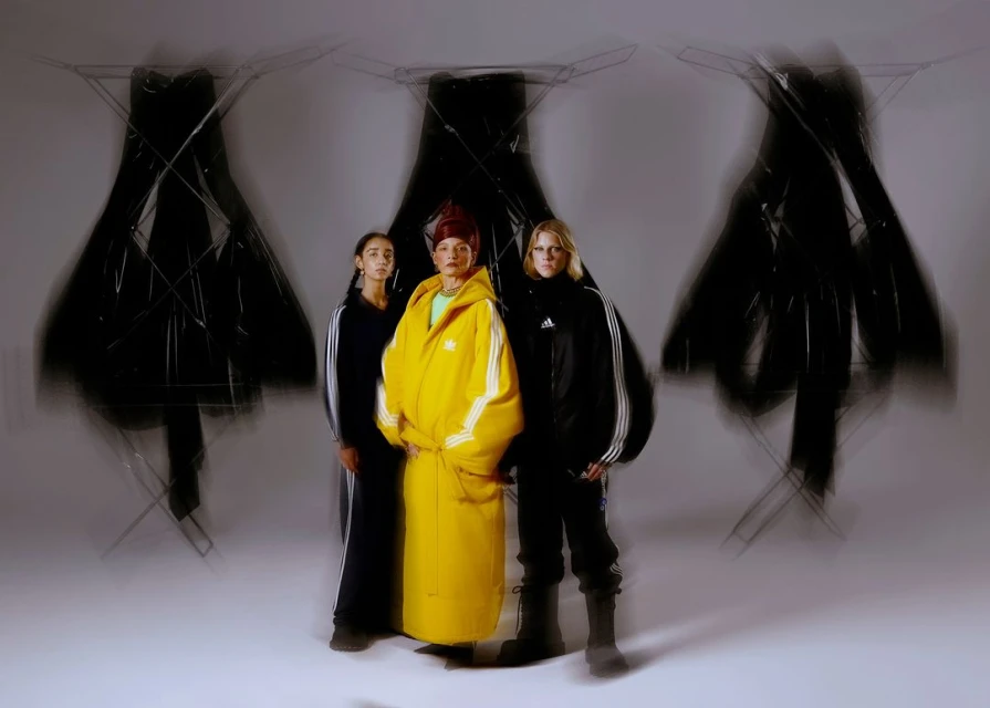 Three people pose in Adidas apparel in front of abstract sculptures. One wears a yellow robe and two others wear black tracksuits.