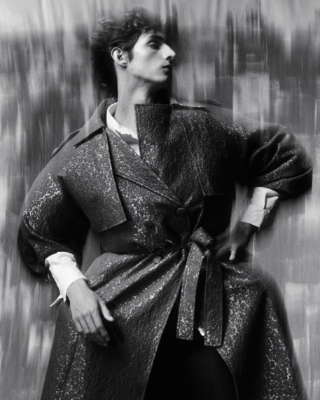 Man wearing a textured belted coat and white shirt posing against a textured background in a black and white portrait.