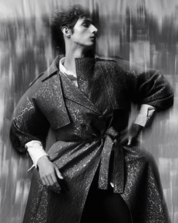 Man wearing a textured belted coat and white shirt posing against a textured background in a black and white portrait.