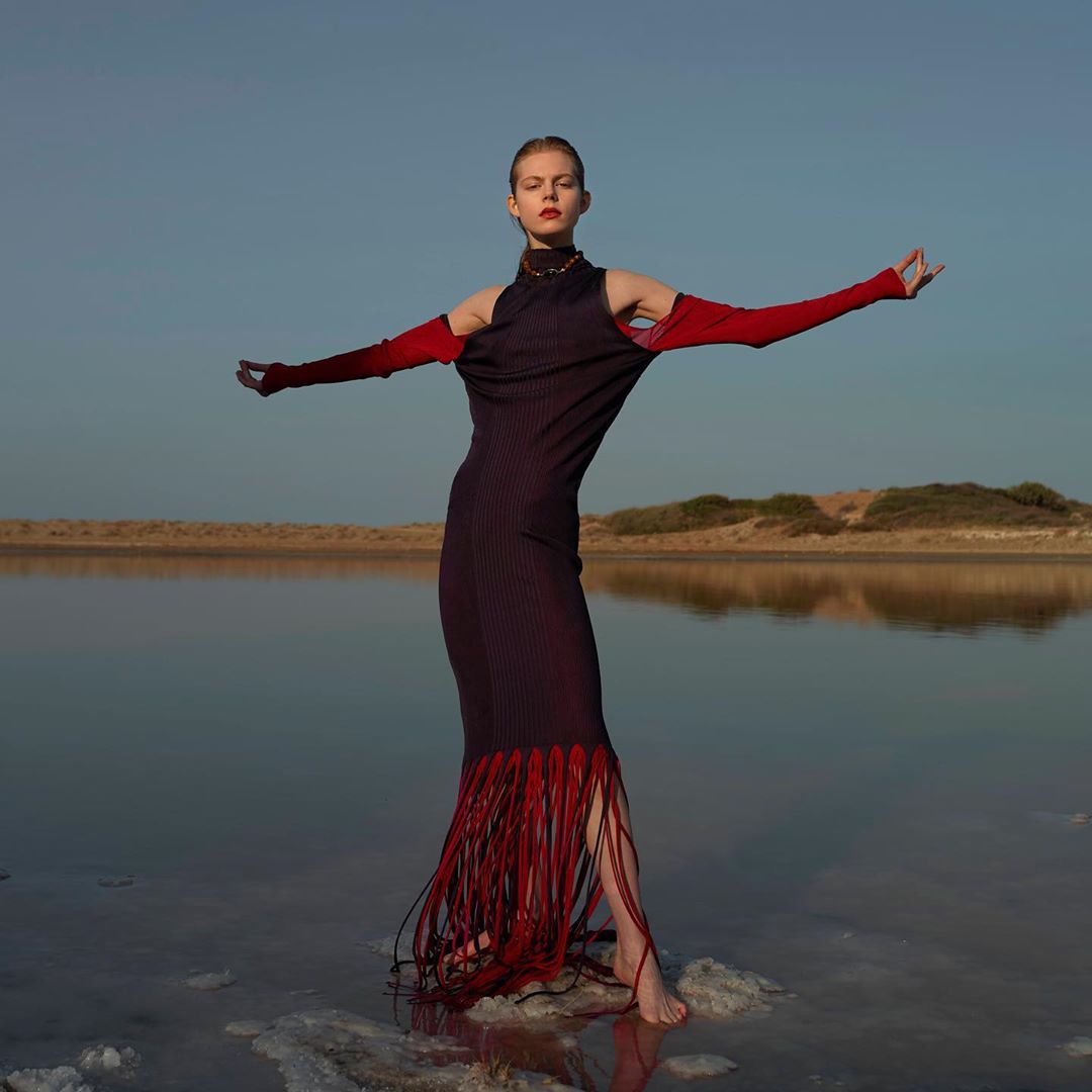 A slender woman with open arms models a dark dress with red fringe at the water's edge under a clear sky during daytime.