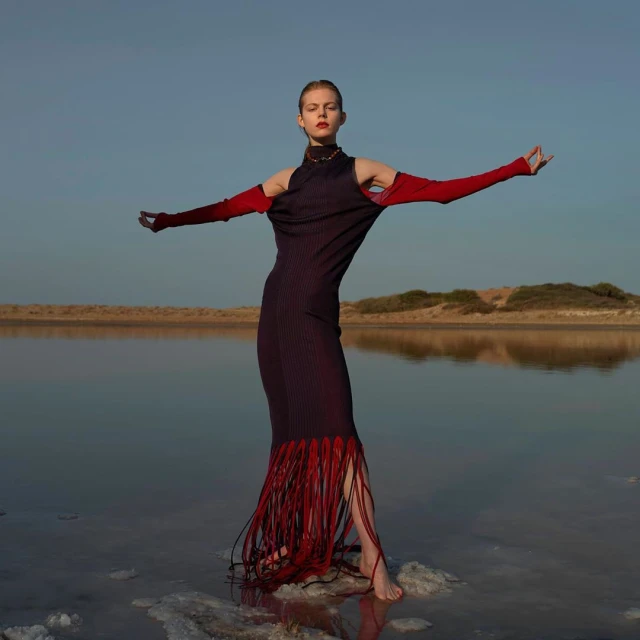 A slender woman with open arms models a dark dress with red fringe at the water's edge under a clear sky during daytime.