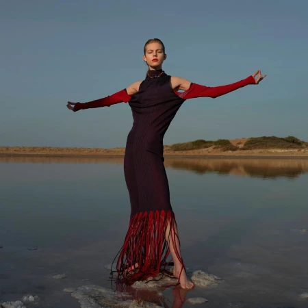 A slender woman with open arms models a dark dress with red fringe at the water's edge under a clear sky during daytime.