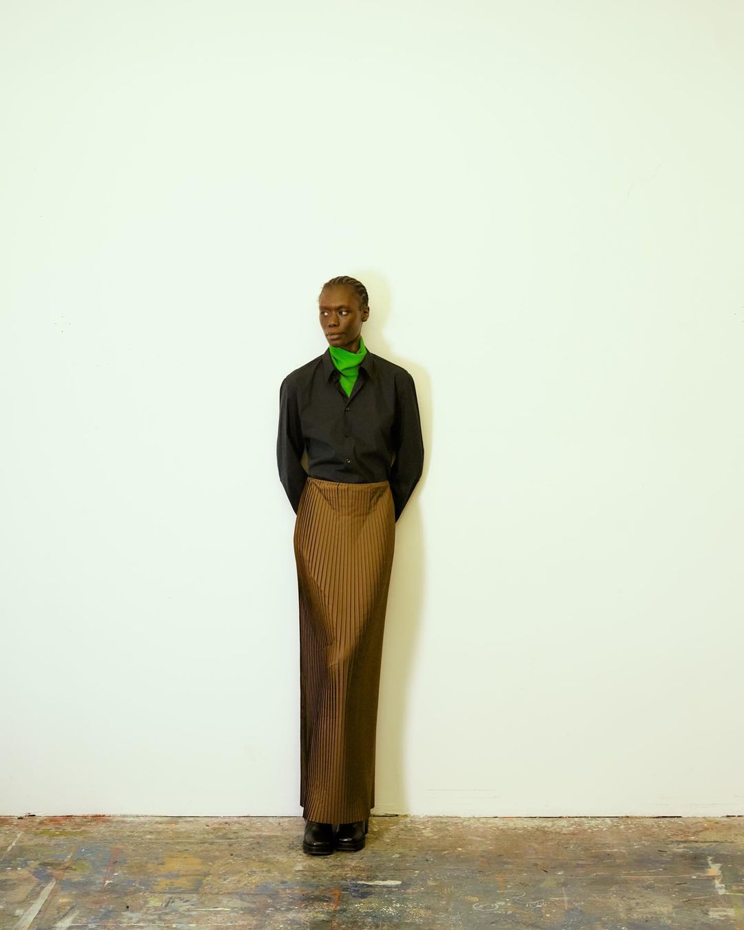 A slender person stands against a white wall wearing a black shirt and a long, pleated brown skirt, hands clasped behind their back.