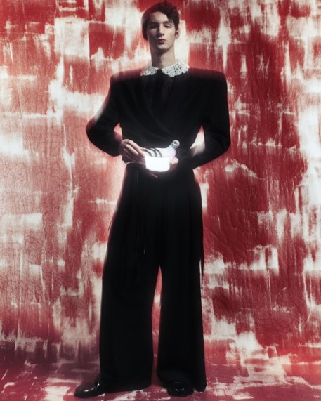 Full-length fashion shot of a young adult male posing in black wrap jacket and wide-leg pants against a textured red and white backdrop.