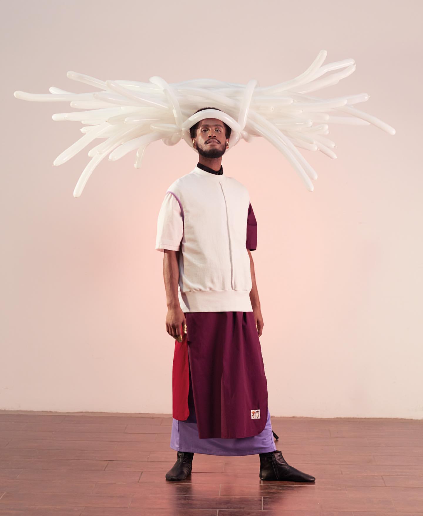 An adult man wearing a hat with white balloon-like ornamentation, a short-sleeved white bomber jacket, and a layered skirt poses indoors.