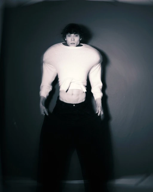 Unique studio portrait of a model wearing a cropped white sweater and dark pants, with muted studio lighting against a gray wall.
