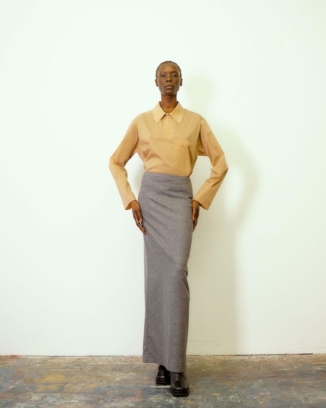 Elegant full shot of a Black female fashion model in tan button-down and a long gray skirt, posing against a white background.