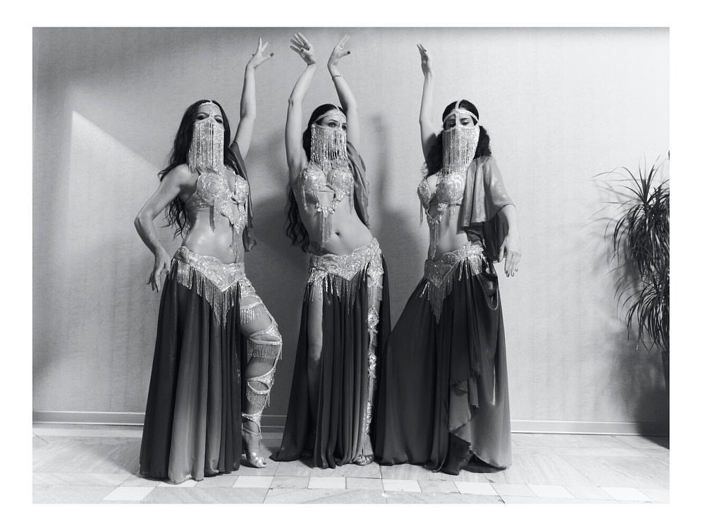 Three belly dancers in traditional attire pose indoors against a textured wall, showcasing elaborate costumes and symmetrical composition.