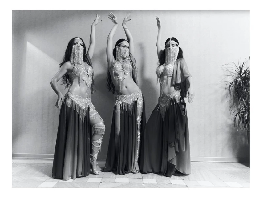 Three belly dancers in traditional attire pose indoors against a textured wall, showcasing elaborate costumes and symmetrical composition.