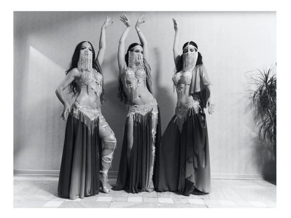 Three belly dancers in traditional attire pose indoors against a textured wall, showcasing elaborate costumes and symmetrical composition.