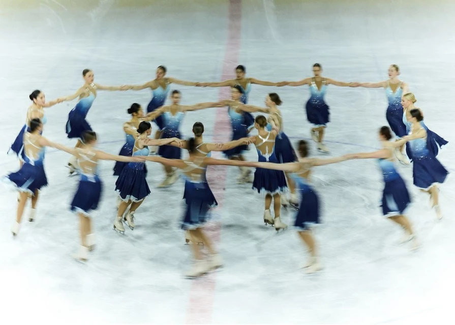 A synchronized ice-skating team of women holding hands in a circle, wearing matching blue dresses on an indoor ice rink.